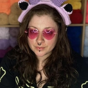 May include: A person wearing a purple crocheted headband with horns and black ears. The headband has a pink band. The person is wearing pink heart-shaped sunglasses and has a nose ring and a lip ring.