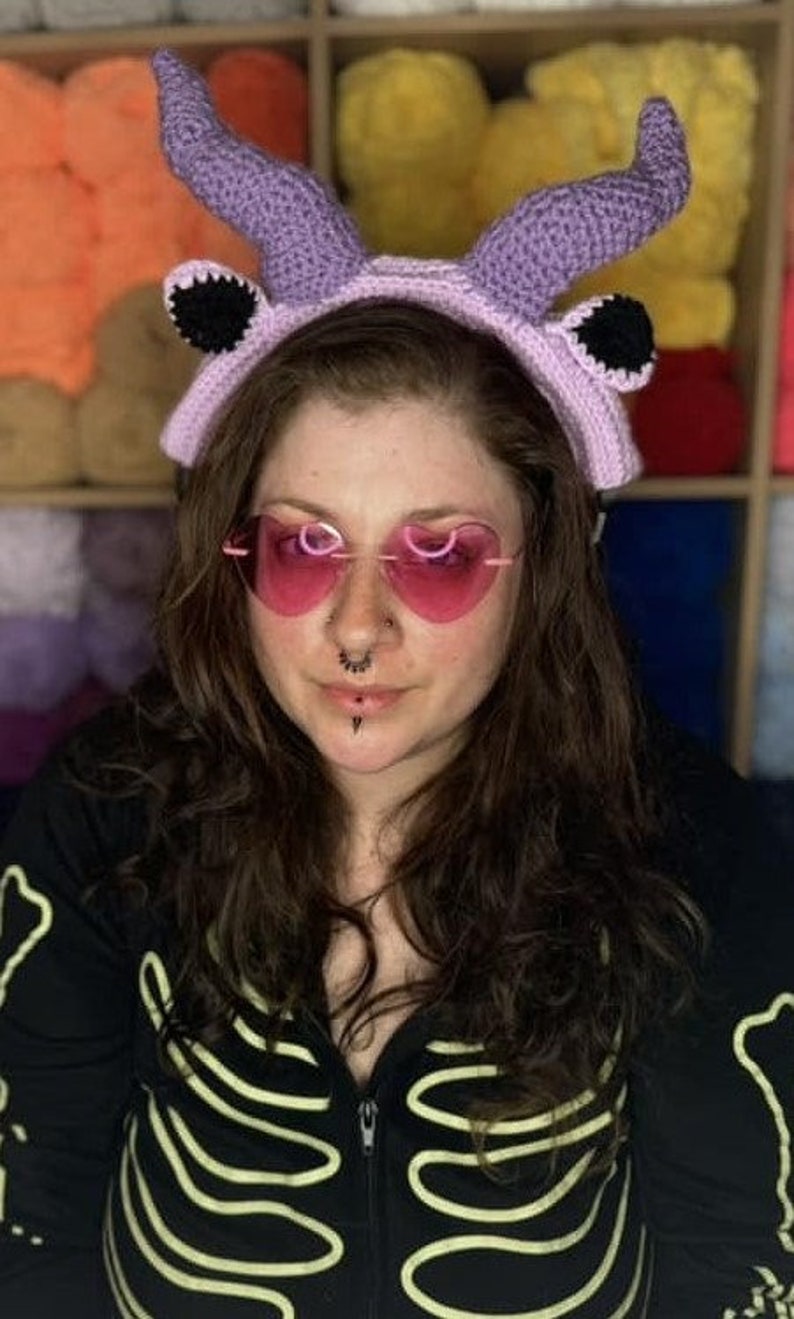 May include: A person wearing a purple crocheted headband with horns and black ears. The headband has a pink band. The person is wearing pink heart-shaped sunglasses and has a nose ring and a lip ring.