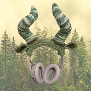 May include: Green and white crocheted headband with large horns, designed to fit over headphones. The headband is in front of a blurred background of green trees.