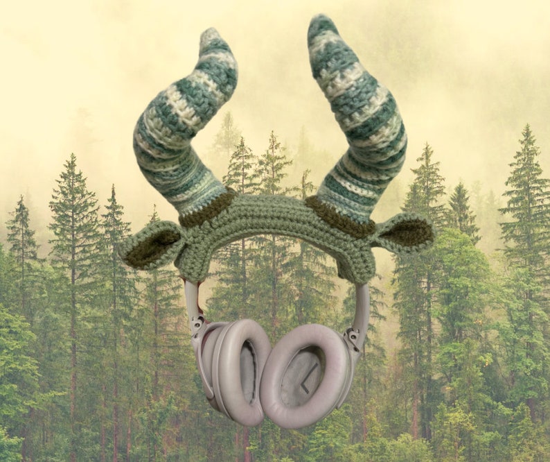 May include: Green and white crocheted headband with large horns, designed to fit over headphones. The headband is in front of a blurred background of green trees.