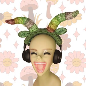 May include: A green and multi-colored crocheted headband with a pair of black headphones. The headband is worn by a mannequin with a bright pink mouth.