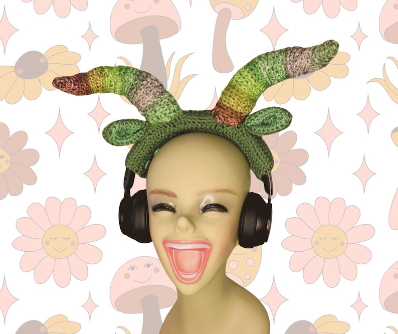 May include: A green and multi-colored crocheted headband with a pair of black headphones. The headband is worn by a mannequin with a bright pink mouth.