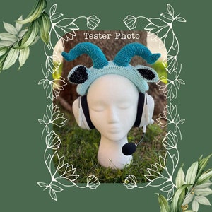 May include: A blue and green crocheted headband with horns, resembling a goat or ram, is worn on a white mannequin head. The headband is paired with white headphones and a microphone.