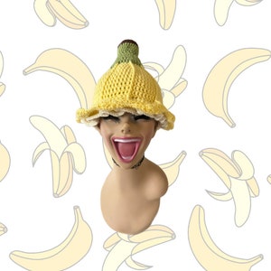 May include: A yellow crochet hat shaped like a banana, with a green stem and brown tip. The hat has a ruffled, cream-colored edge. The background features a pattern of banana illustrations.