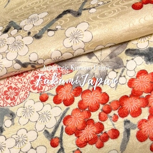 Può includere: Un tessuto dorato con un motivo di fiori di prugno bianchi e rossi su rami scuri, con un sottile sfondo a onde e accenti floreali geometrici rossi. Il testo Authentic Kimono Fabric FukumiJapan è stampato in bianco al centro.