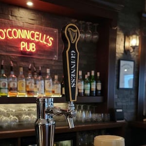 Op de afbeelding: Een bar interieur met een Guinness tapkraan, met een harp logo en het woord "Guinness". Het neonbord erboven luidt "O'Connell's Pub". Verschillende flessen sterke drank zijn zichtbaar op planken op de achtergrond.