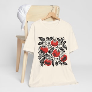 May include: A cream-colored t-shirt with a black and red illustration of tomatoes on a vine. The illustration is centered on the shirt.