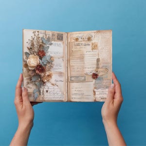 May include: An open vintage scrapbook with aged paper pages, adorned with dried flowers, stamps, and ephemera. The left side features a floral arrangement with cream, burgundy, and blue-grey accents. The background is a solid blue.