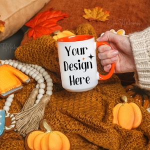 Puede incluir: Taza de cerámica blanca con borde y asa naranja. La taza tiene el texto "Your Design Here" impreso en ella. La taza está sobre una manta de punto marrón con decoraciones de otoño.