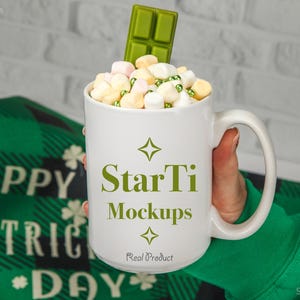 May include: White ceramic mug filled with marshmallows and green sprinkles, topped with a green chocolate bar. The mug has the text "StarTi Mockups" and "Real Product" printed on it. The mug is being held by a hand wearing a green sweater.