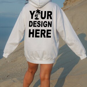 Puede incluir: Sudadera con capucha blanca con el texto "YOUR DESIGN HERE" y un gr&aacute;fico de palmera impreso en la espalda. La sudadera la lleva puesta una persona que camina por una playa de arena.
