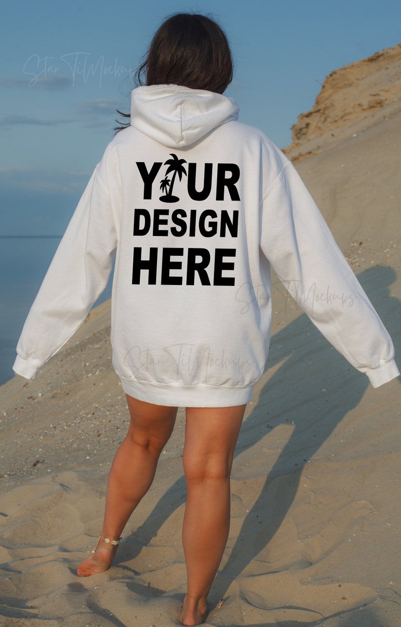 Puede incluir: Sudadera con capucha blanca con el texto "YOUR DESIGN HERE" y un gr&aacute;fico de palmera impreso en la espalda. La sudadera la lleva puesta una persona que camina por una playa de arena.