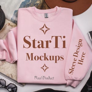 Pu&ograve; includere: Una felpa rosa con il testo "StarTi Mockups" e "Sleeve Design Here" stampato su di essa. La felpa &egrave; stesa su una superficie bianca con altri oggetti intorno.