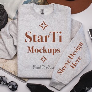 Pu&ograve; includere: Una felpa grigia con il testo "StarTi Mockups" e "Sleeve Design Here" stampato su di essa. La felpa &egrave; stesa su una superficie bianca con altri oggetti intorno.