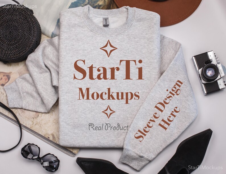 Pu&ograve; includere: Una felpa grigia con il testo "StarTi Mockups" e "Sleeve Design Here" stampato su di essa. La felpa &egrave; stesa su una superficie bianca con altri oggetti intorno.