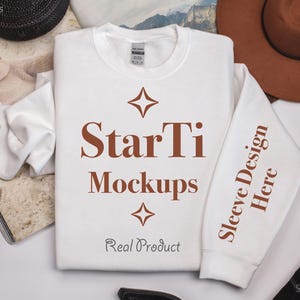 Pu&ograve; includere: Felpa bianca a maniche lunghe con un design di testo marrone che dice "StarTi Mockups" e "Sleeve Design Here" sulla manica. Il testo &egrave; centrato sulla felpa e ha un design a stella sopra e sotto il testo. Il testo "Real Product" &egrave; sotto il design a stella.