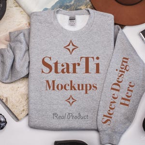 Pu&ograve; includere: Una felpa grigia con il testo "StarTi Mockups" e "Sleeve Design Here" stampato su di essa. La felpa &egrave; stesa su una superficie bianca con altri oggetti intorno.