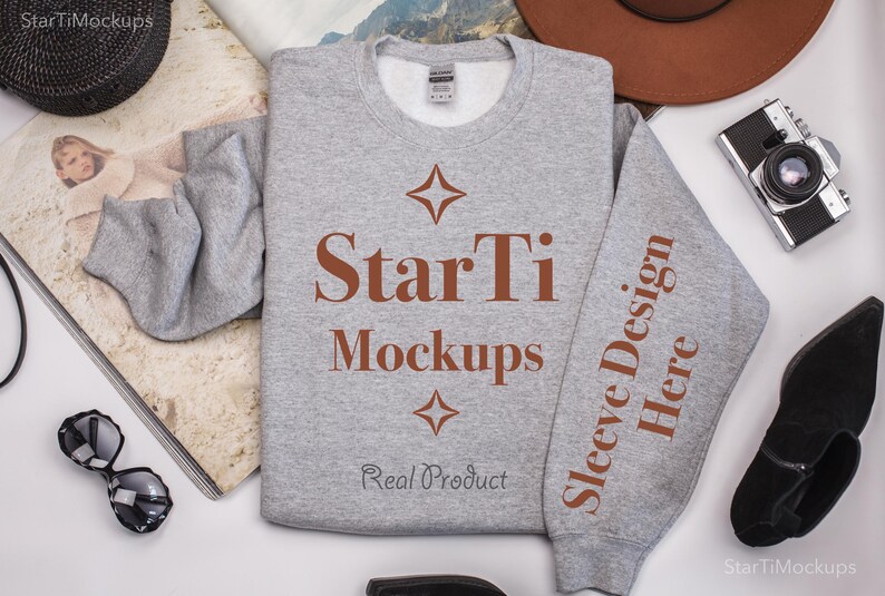 Pu&ograve; includere: Una felpa grigia con il testo "StarTi Mockups" e "Sleeve Design Here" stampato su di essa. La felpa &egrave; stesa su una superficie bianca con altri oggetti intorno.