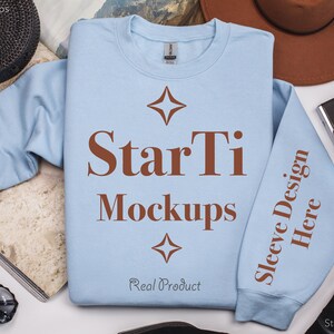 Pu&ograve; includere: Una felpa di colore azzurro chiaro con il testo "StarTi Mockups" e "Sleeve Design Here" stampato in marrone. La felpa &egrave; appoggiata su una superficie bianca con altri oggetti intorno.