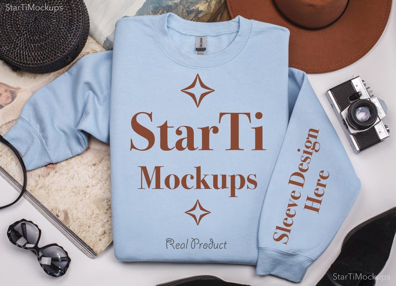 Pu&ograve; includere: Una felpa di colore azzurro chiaro con il testo "StarTi Mockups" e "Sleeve Design Here" stampato in marrone. La felpa &egrave; appoggiata su una superficie bianca con altri oggetti intorno.