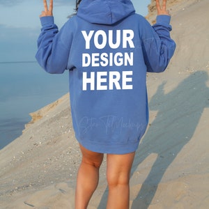 Puede incluir: Una mujer con una sudadera con capucha azul claro con el texto "YOUR DESIGN HERE" impreso en la espalda. Est&aacute; de pie en una playa de arena de espaldas a la c&aacute;mara.