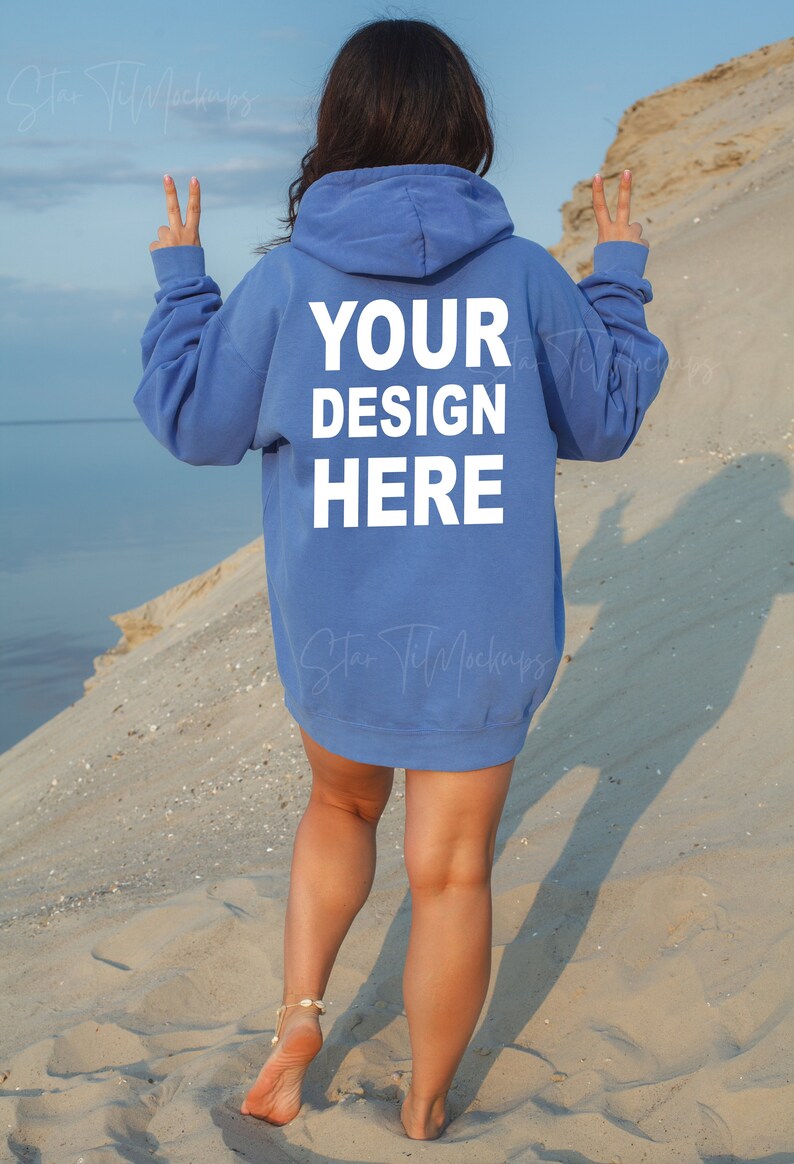Puede incluir: Una mujer con una sudadera con capucha azul claro con el texto "YOUR DESIGN HERE" impreso en la espalda. Est&aacute; de pie en una playa de arena de espaldas a la c&aacute;mara.