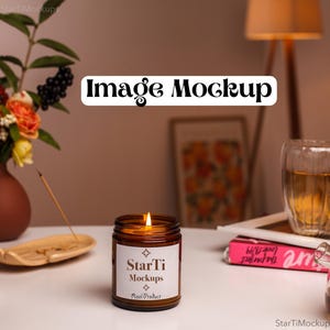 May include: A brown glass candle jar with a white label that reads "StarTi Mockups Real Product" and a lit candle inside. The candle is on a white table with a wooden incense holder and a pink book.