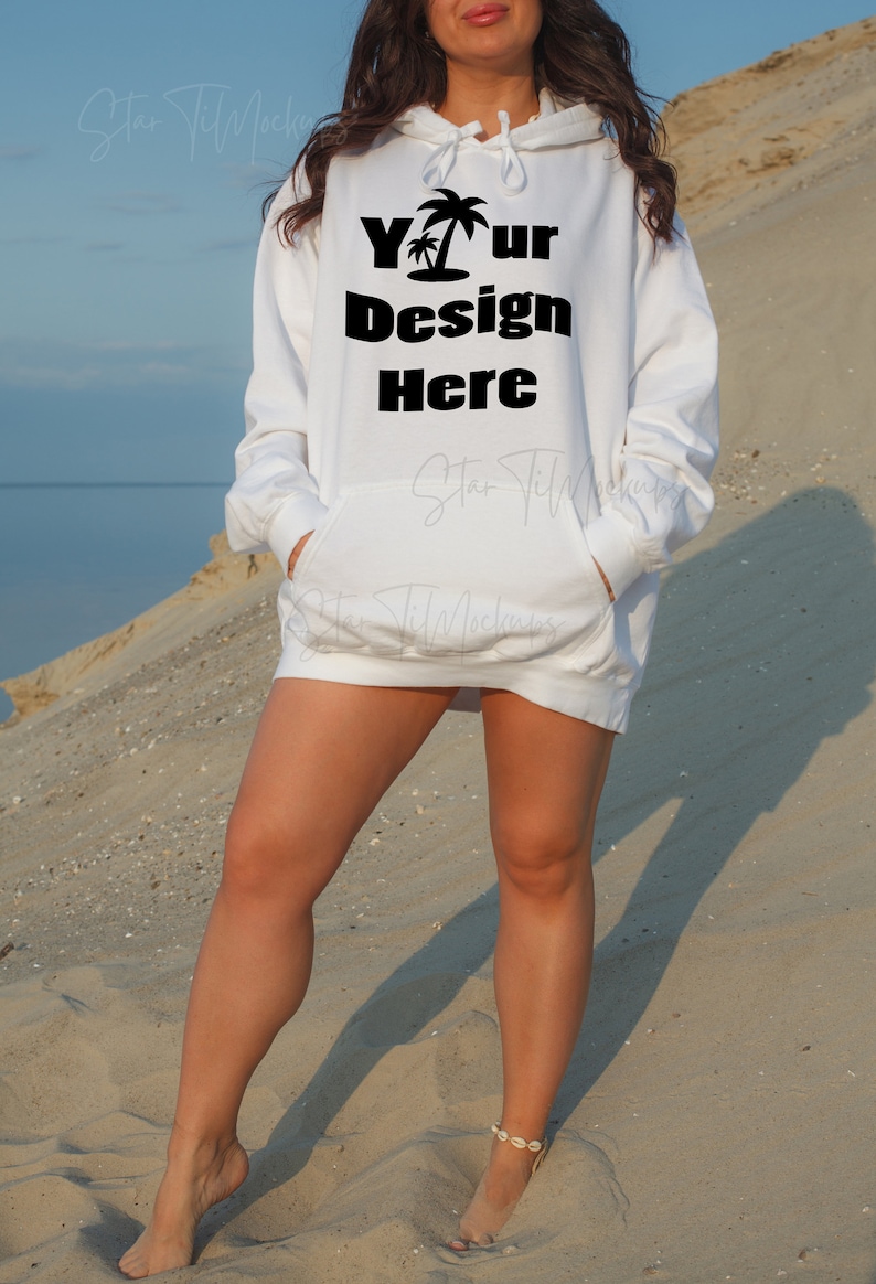 Puede incluir: Una sudadera con capucha blanca con el texto "Your Design Here" y un gr&aacute;fico de palmera impreso en la parte delantera. La sudadera la lleva puesta una persona de pie en una playa de arena.