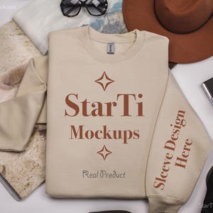 Pu&ograve; includere: Una felpa beige a maniche lunghe e girocollo. La felpa ha il testo "StarTi Mockups" e "Real Product" stampato sul davanti. Il testo "Sleeve Design Here" &egrave; stampato sulla manica destra.