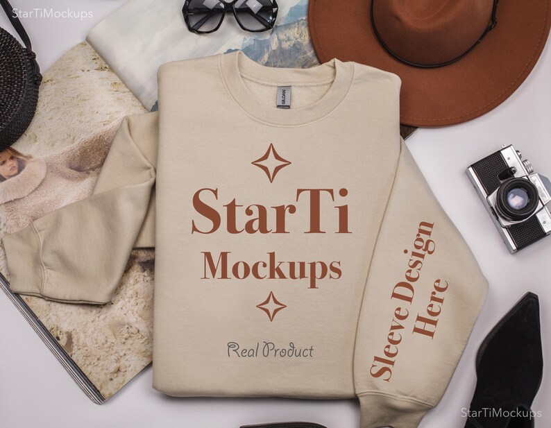 Pu&ograve; includere: Una felpa beige a maniche lunghe e girocollo. La felpa ha il testo "StarTi Mockups" e "Real Product" stampato sul davanti. Il testo "Sleeve Design Here" &egrave; stampato sulla manica destra.