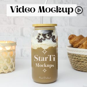 Puede incluir: Un tarro de vidrio con tapa marrón y una pajita, lleno de una bebida de café cubierta con crema batida y virutas de chocolate. El tarro tiene el texto "StarTi Mockups" en él. El tarro está sobre una superficie blanca con una cesta de mimbre de croissants en el fondo.