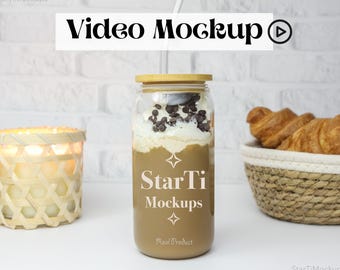 May include: A glass jar with a brown lid and a straw, filled with a coffee drink topped with whipped cream and chocolate chips. The jar has the text "StarTi Mockups" on it. The jar is on a white surface with a wicker basket of croissants in the background.