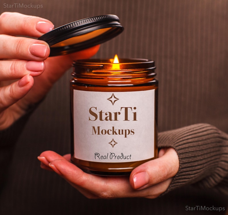 May include: A lit brown glass candle jar with a black lid is held in a person's hands. The candle has a white label with the text "StarTi Mockups" and "Real Product". The candle is lit with a bright flame.