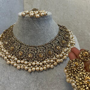 May include: A gold-toned necklace and earring set with faux pearls and decorative accents. The necklace is displayed on a gray bust, and the earrings are held in a hand. The set features a detailed design with a vintage aesthetic.