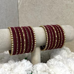 Könnte beinhalten: Zwei Sets aus bordeauxroten Samtarmreifen mit goldfarbenen Metallakzenten und kleinen weißen Kunstperlen. Jedes Set besteht aus mehreren dünnen Armreifen, die zusammen angeordnet sind und eine strukturierte Oberfläche aufweisen.