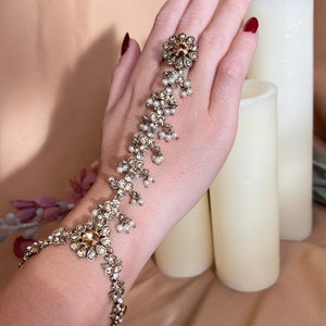 May include: A gold hand and finger jewellery piece with white pearls and clear rhinestones. The jewellery is worn on a hand with red nail polish.