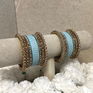 May include: Two bangles featuring a central light blue band with small gold dots, flanked by multiple thin gold-toned textured bands. The bangles are constructed with a combination of smooth and beaded metallic elements, creating a layered appearance.