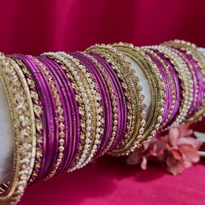 Könnte beinhalten: Ein Set aus Armreifen mit abwechselnden Bändern aus metallischem Gold und leuchtendem Magenta. Die Kollektion umfasst schlichte magentafarbene Ringe, strukturierte Goldbänder und dünne Armreifen mit kleinen weißen Perlen in gestapelter Form.