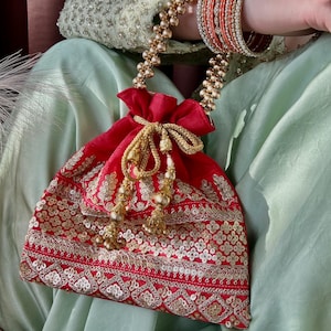 Może przedstawiać: Czerwona torba ze sznurkiem, złotym haftem i złotym uchwytem z koralików. Torba jest ozdobiona cekinami i frędzlami. Na nadgarstku stos pomarańczowych i złotych bransoletek.