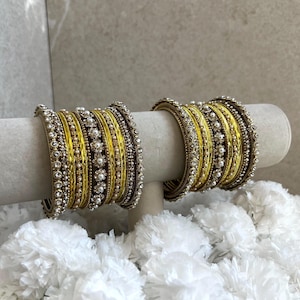 May include: Two sets of stacked bangles featuring alternating bands of bright yellow metallic finish and rows of small white faux pearls. Each bangle set is composed of multiple thin rings with intricate beaded textures and clear crystal accents.