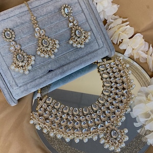 May include: A gold-toned jewelry set featuring a necklace, earrings, and a headpiece. The pieces are adorned with clear, teardrop-shaped stones and small, white pearls. The set is displayed on a reflective surface with white floral accents.