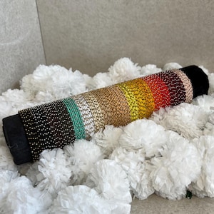 May include: A cylindrical beaded bracelet featuring a gradient of colors including black, teal, white, gold, yellow, orange, and red. The beads are arranged in horizontal rows around the black fabric core, creating a vibrant, multi-colored pattern.