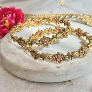 Puede incluir: Dos brazaletes de oro con intrincados diseños florales y piedras preciosas rosas. Los brazaletes están apilados uno encima del otro.