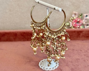 May include: Gold hoop earrings with dangling pearl and bead embellishments. The earrings are displayed on a white stand.