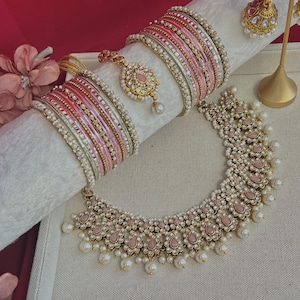 May include: A jewellery set in pink and white, including a necklace, earrings, bangles, and a headpiece. The necklace and headpiece have pink accents and pearl-like beads. The bangles are a mix of pink, white, and gold. The earrings are gold with pink and white details.