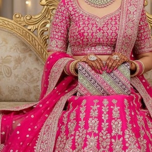 May include: A woman in a vibrant pink embroidered lehenga, a traditional Indian outfit, sits on a gold ornate sofa. The outfit includes a matching blouse, skirt, and dupatta, all adorned with intricate silver embroidery. She holds a patterned clutch.