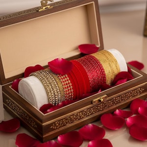 May include: A rectangular wooden jewelry box with a hinged lid and gold-tone latch, containing a white velvet cylinder display stand. The stand holds a collection of stacked bangles in alternating red, gold, and crystal-encrusted textures.