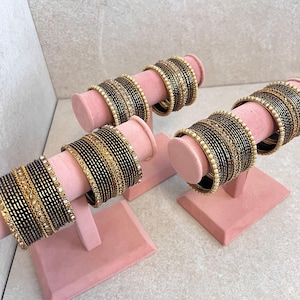 May include: Several stacks of black and gold bangles are displayed on pink velvet stands. The bangles have a mix of solid black bands and bands adorned with gold and clear gemstones. The stands are square-based with a cylindrical top.