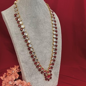 Peut inclure: Un collier doré orné de pierres précieuses claires et rouges. Le collier est présenté sur un buste gris sur fond rouge. Le collier présente un motif répété de pierres claires et facettées et de petites perles rouges.