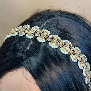 Może przedstawiać: Złota opaska do włosów z białymi perełkami i przezroczystymi cyrkoniami. Opaska ozdobiona jest misternymi wzorami kwiatowymi.